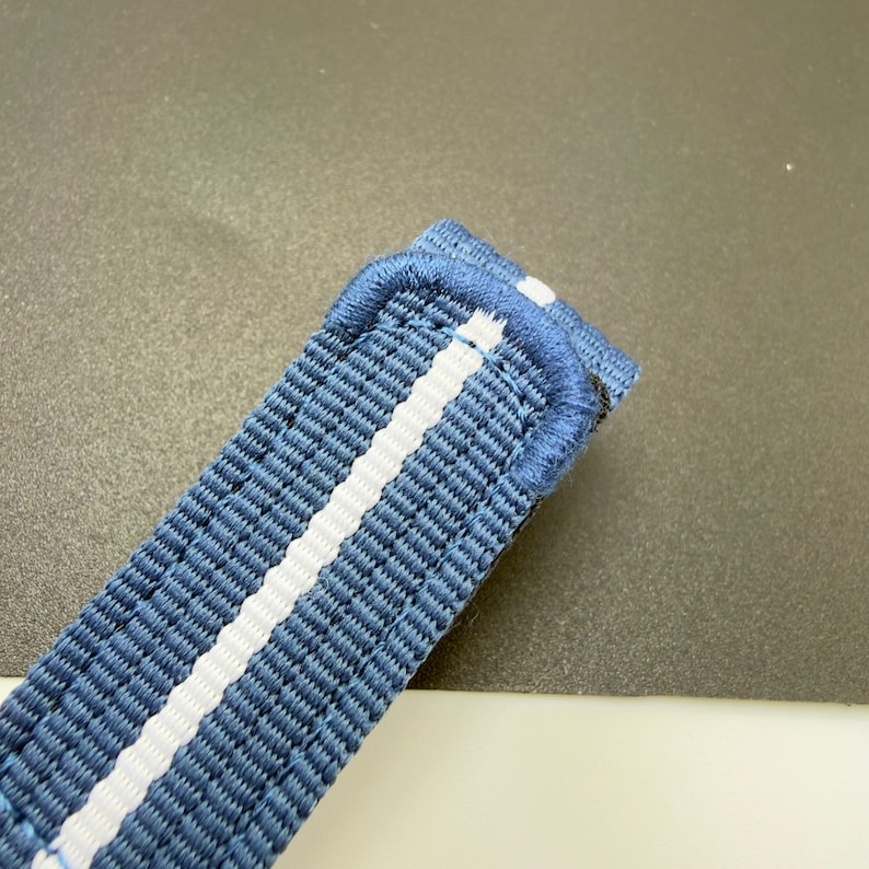 Puede incluir: Una correa de reloj tejida en color azul con una &uacute;nica franja vertical blanca en el centro. La correa presenta un borde cosido reforzado y una superficie de tela texturizada, mostrando una construcci&oacute;n duradera con una trabilla.