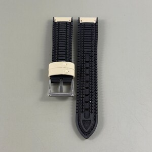 May include: A black watch strap with a textured pattern. The strap has a cream-colored band and a silver buckle. The strap is designed to be durable and stylish.