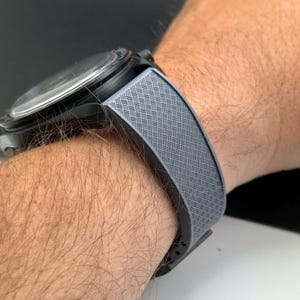 May include: A close-up of a black wristwatch with a gray patterned band. The band has a woven design. The watch face is black with a silver-colored bezel. The watch is on a person's wrist.