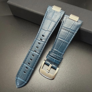 May include: Two blue watch straps with a crocodile skin texture. The straps have silver-colored metal clasps and buckles. The straps are made of a durable material and are designed to attach to a watch face. The straps are displayed on a dark surface.