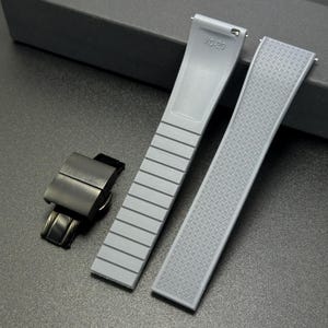 May include: A gray watch strap set with a black clasp. The strap has a textured pattern on one side and a ribbed design on the other. The strap is marked with "XO-20".