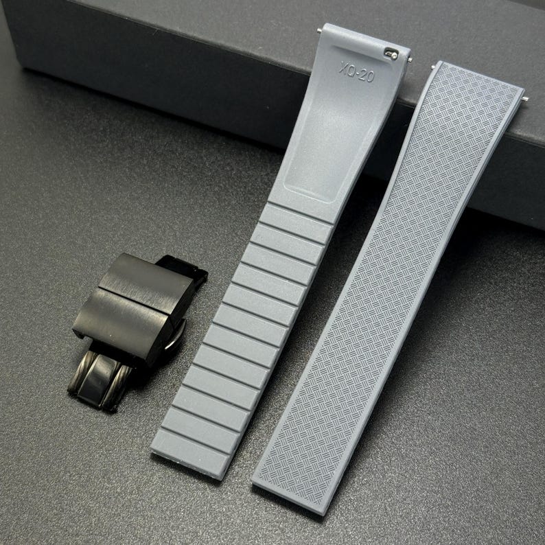 May include: A gray watch strap set with a black clasp. The strap has a textured pattern on one side and a ribbed design on the other. The strap is marked with "XO-20".