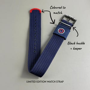Puede incluir: Una correa de reloj tejida en azul marino con punta roja, hebilla y trabilla negras, y un emblema bordado en rojo, blanco y azul en la trabilla. El texto dice Coloured to match, Black buckle + keeper y LIMITED EDITION WATCH STRAP.