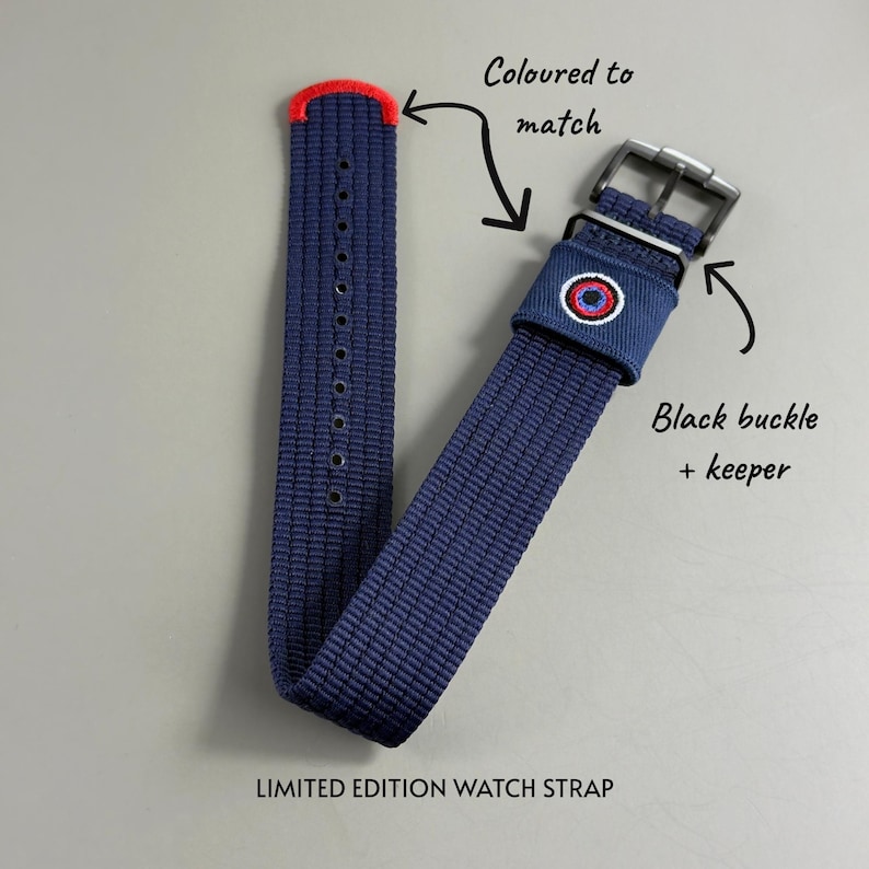 Puede incluir: Una correa de reloj tejida en azul marino con punta roja, hebilla y trabilla negras, y un emblema bordado en rojo, blanco y azul en la trabilla. El texto dice Coloured to match, Black buckle + keeper y LIMITED EDITION WATCH STRAP.