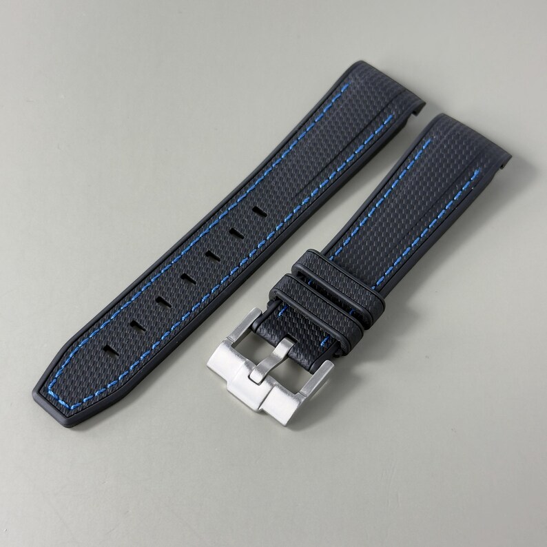 Puede incluir: Una correa de reloj negra con una superficie texturizada y costuras azules a lo largo de los bordes. La correa incluye una hebilla de metal plateado y dos trabillas negras a juego para asegurar la banda, dise&ntilde;ada para una caja de reloj.