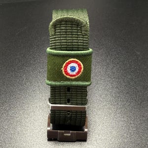 Puede incluir: Una correa de reloj de nailon verde oliva con textura tejida y hebilla de metal. Un peque&ntilde;o parche circular bordado est&aacute; centrado en la correa, mostrando un dise&ntilde;o de escarapela roja, blanca y azul con un borde de color dorado.