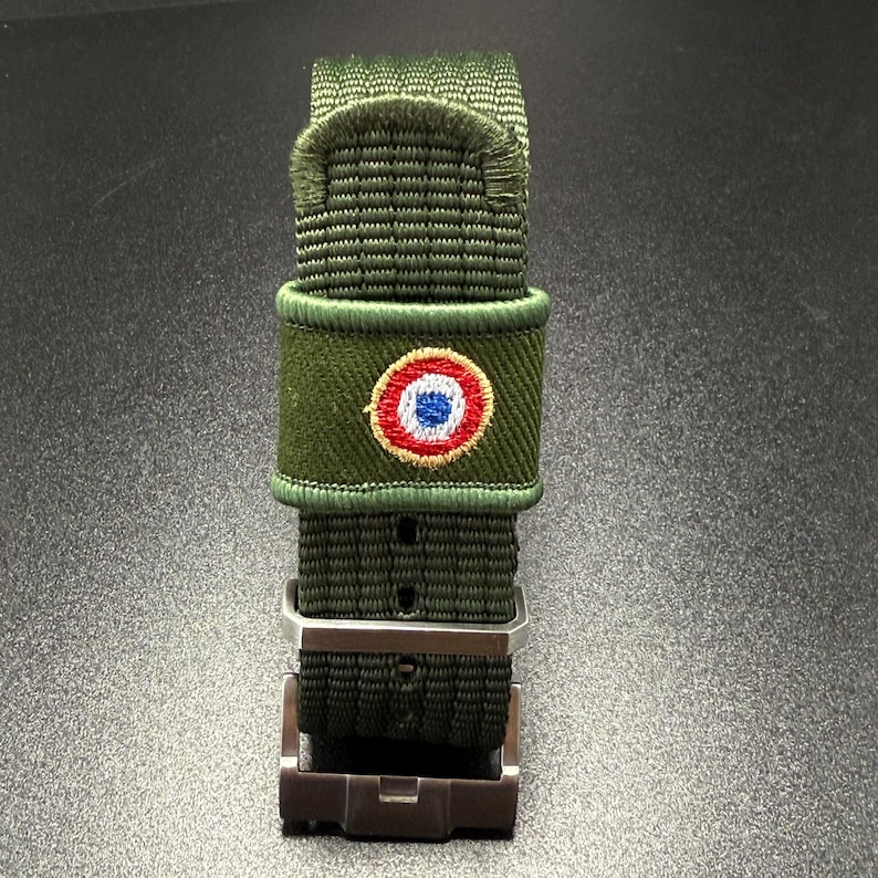 Puede incluir: Una correa de reloj de nailon verde oliva con textura tejida y hebilla de metal. Un peque&ntilde;o parche circular bordado est&aacute; centrado en la correa, mostrando un dise&ntilde;o de escarapela roja, blanca y azul con un borde de color dorado.