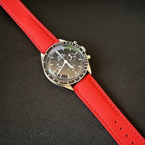 Puede incluir: Un reloj de pulsera con esfera negra, bisel plateado y correa roja. La esfera del reloj presenta m&uacute;ltiples subesferas y una escala taquim&eacute;trica. La correa roja es de material tejido y est&aacute; cosida con hilo en contraste.