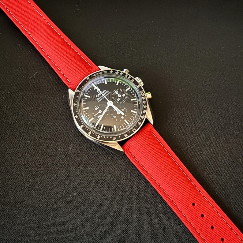 Puede incluir: Un reloj de pulsera con esfera negra, bisel plateado y correa roja. La esfera del reloj presenta m&uacute;ltiples subesferas y una escala taquim&eacute;trica. La correa roja es de material tejido y est&aacute; cosida con hilo en contraste.