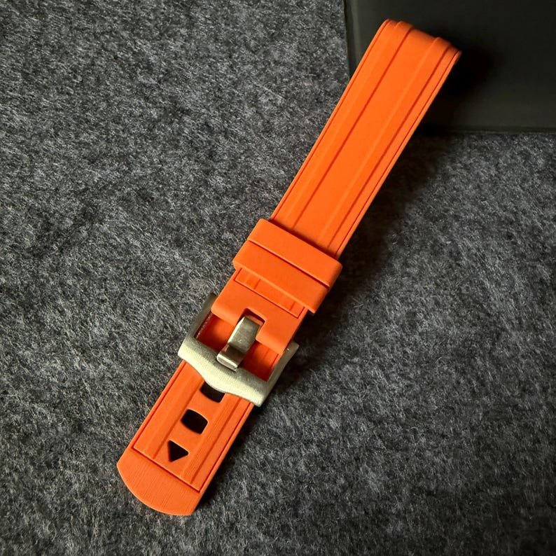 Puede incluir: Correa de reloj naranja brillante con hebilla plateada. La correa est&aacute; hecha de un material flexible y acanalado. La hebilla tiene forma rectangular con cierre de pasador. La correa mide aproximadamente 23 cm de largo.