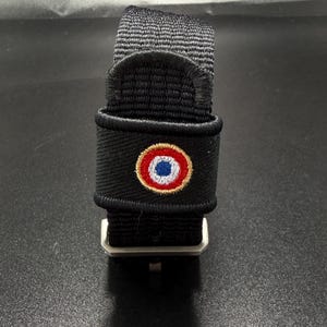 Puede incluir: Una correa de reloj de nailon negro con un emblema circular bordado que presenta anillos conc&eacute;ntricos en azul, blanco y rojo, bordeado por un hilo de color dorado. La correa incluye una hebilla met&aacute;lica y un acabado de tela texturizada.