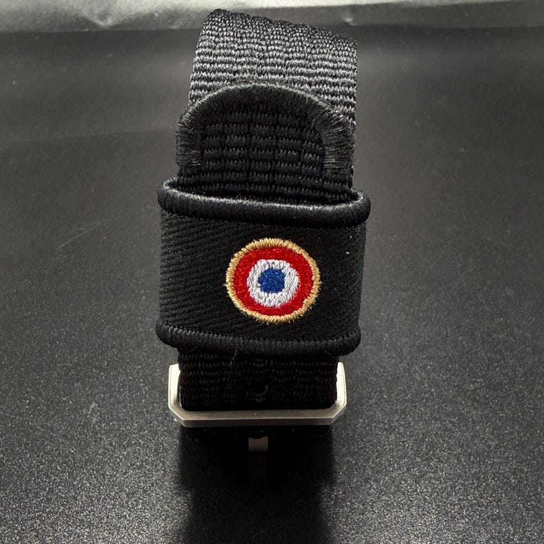 Puede incluir: Una correa de reloj de nailon negro con un emblema circular bordado que presenta anillos conc&eacute;ntricos en azul, blanco y rojo, bordeado por un hilo de color dorado. La correa incluye una hebilla met&aacute;lica y un acabado de tela texturizada.