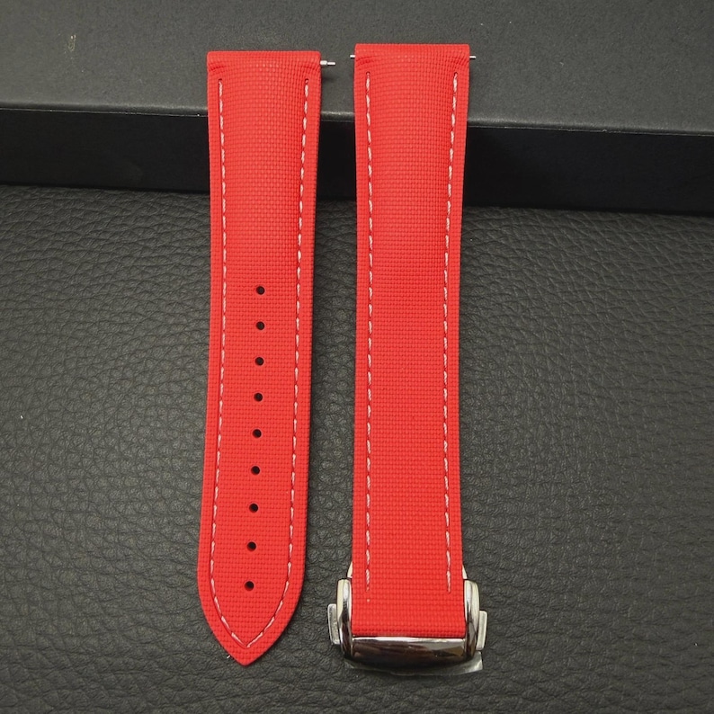 Puede incluir: Correa de reloj roja brillante con costuras blancas. La correa tiene un cierre de acero inoxidable y m&uacute;ltiples orificios de ajuste. La correa del reloj est&aacute; hecha de un material texturizado.