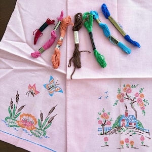 May include: Two pink embroidered tea towels with floral and butterfly designs, alongside spools of colorful embroidery floss. One towel features a house and tree scene, the other has butterflies and water plants.