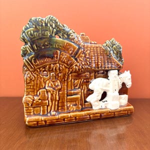 May include: A ceramic figurine depicting a rustic scene with a thatched roof building, a figure, and a horse-drawn cart. The figurine is primarily brown with green and white accents. The text "Under the Spread Chestnut" is visible.