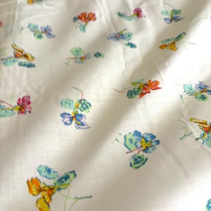 May include: White fabric with a repeating pattern of colorful floral designs. The flowers are in shades of blue, orange, yellow, and pink, with green leaves. The fabric appears to be lightweight and slightly wrinkled.