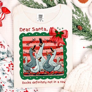 Puede incluir: Sudadera color crema con un gr&aacute;fico de patos de dibujos animados graznando. El dise&ntilde;o incluye el texto "Dear Santa," y "Ducks definitely not in a row." Un lazo rojo y un borde verde enmarcan los gansos.