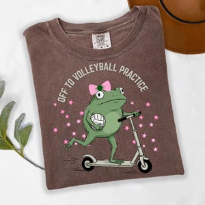 May include: Brown t-shirt with the text "OFF TO VOLLEYBALL PRACTICE". The shirt features a cartoon frog riding a scooter and holding a volleyball. The frog wears a pink bow and is surrounded by pink stars.