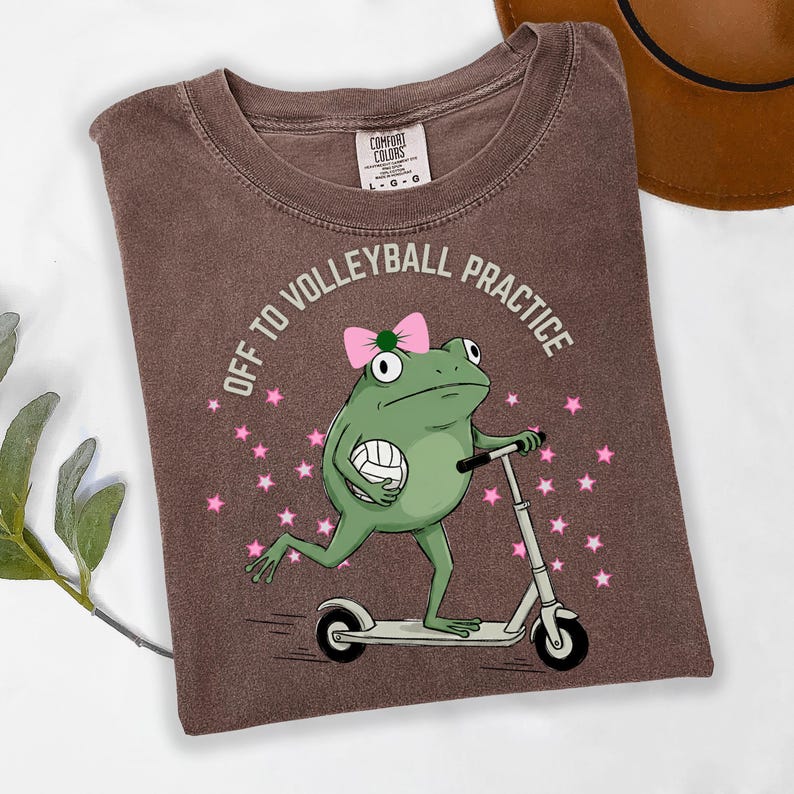 May include: Brown t-shirt with the text "OFF TO VOLLEYBALL PRACTICE". The shirt features a cartoon frog riding a scooter and holding a volleyball. The frog wears a pink bow and is surrounded by pink stars.