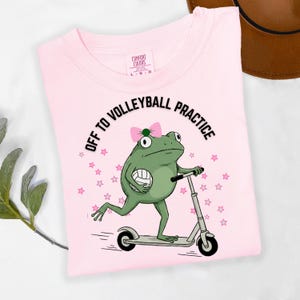 May include: A light pink t-shirt with the text "OFF TO VOLLEYBALL PRACTICE" and a cartoon frog riding a scooter while holding a volleyball. The frog has a pink bow and is surrounded by pink stars. The shirt is made of Comfort Colors material.