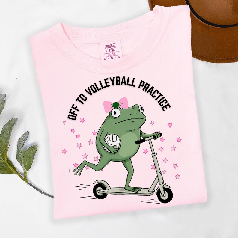 May include: A light pink t-shirt with the text "OFF TO VOLLEYBALL PRACTICE" and a cartoon frog riding a scooter while holding a volleyball. The frog has a pink bow and is surrounded by pink stars. The shirt is made of Comfort Colors material.