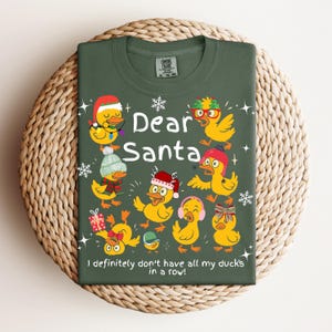 May include: Green t-shirt with the text "Dear Santa" and several cartoon ducks wearing festive accessories. The phrase "I definitely don't have all my ducks in a row!" is printed at the bottom. The shirt is displayed on a woven surface.