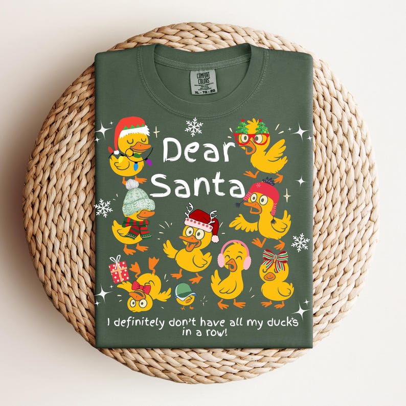 May include: Green t-shirt with the text "Dear Santa" and several cartoon ducks wearing festive accessories. The phrase "I definitely don't have all my ducks in a row!" is printed at the bottom. The shirt is displayed on a woven surface.