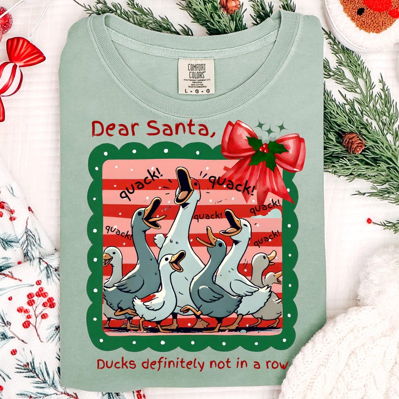 Puede incluir: Camiseta Comfort Colors verde claro con un dise&ntilde;o de patos de dibujos animados y el texto "Dear Santa, Ducks definitely not in a row." El dise&ntilde;o incluye un lazo rojo y la palabra "quack!"