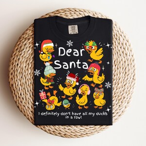 May include: Black t-shirt featuring a festive design with cartoon ducks wearing holiday accessories. The text "Dear Santa" is displayed above the ducks, with the phrase "I definitely don't have all my ducks in a row!" below.