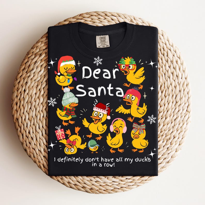 May include: Black t-shirt featuring a festive design with cartoon ducks wearing holiday accessories. The text "Dear Santa" is displayed above the ducks, with the phrase "I definitely don't have all my ducks in a row!" below.
