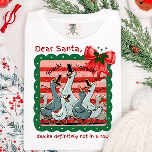 Puede incluir: Camiseta blanca con un gr&aacute;fico de patos de dibujos animados, un lazo rojo y el texto "Dear Santa, Ducks definitely not in a row." Los patos son en tonos grises y blancos.