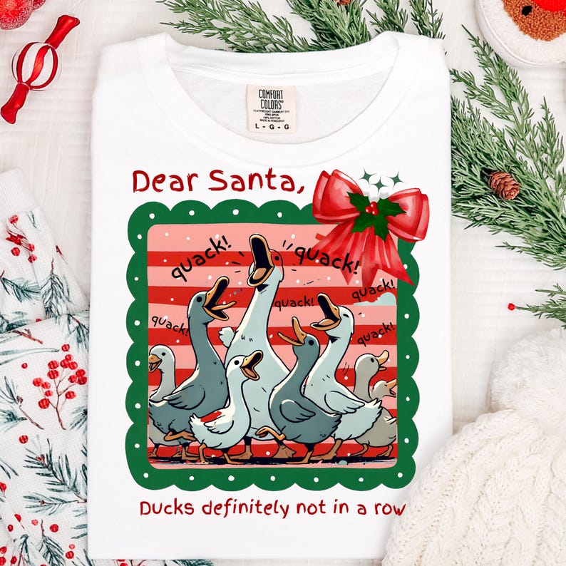 Puede incluir: Camiseta blanca con un gr&aacute;fico de patos de dibujos animados, un lazo rojo y el texto "Dear Santa, Ducks definitely not in a row." Los patos son en tonos grises y blancos.