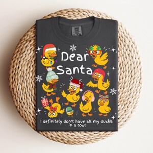 May include: Dark gray t-shirt featuring the text "Dear Santa" with illustrations of yellow ducks wearing festive hats and accessories. The phrase "I definitely don't have all my ducks in a row!" is also printed on the shirt.