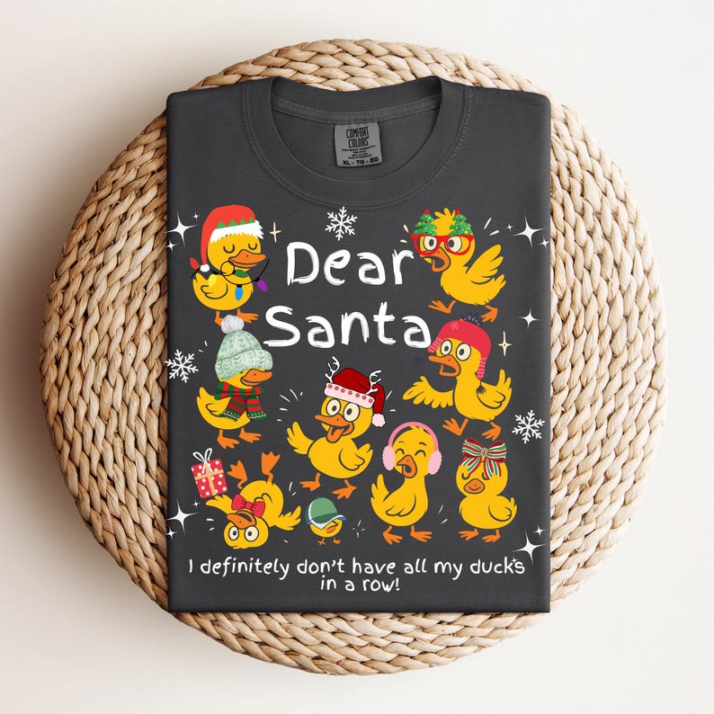 May include: Dark gray t-shirt featuring the text "Dear Santa" with illustrations of yellow ducks wearing festive hats and accessories. The phrase "I definitely don't have all my ducks in a row!" is also printed on the shirt.