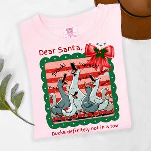 Puede incluir: Camiseta rosa claro con un gr&aacute;fico de patos de dibujos animados. El gr&aacute;fico incluye patos con picos abiertos, la palabra "quack!" y un lazo rojo. El texto dice "Dear Santa, Ducks definitely not in a row".