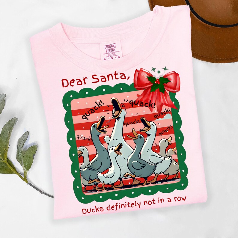 Puede incluir: Camiseta rosa claro con un gr&aacute;fico de patos de dibujos animados. El gr&aacute;fico incluye patos con picos abiertos, la palabra "quack!" y un lazo rojo. El texto dice "Dear Santa, Ducks definitely not in a row".