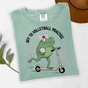 May include: A light green t-shirt with the text "OFF TO VOLLEYBALL PRACTICE". The shirt features a cartoon frog riding a scooter and holding a volleyball. The frog wears a pink bow and is surrounded by pink stars.
