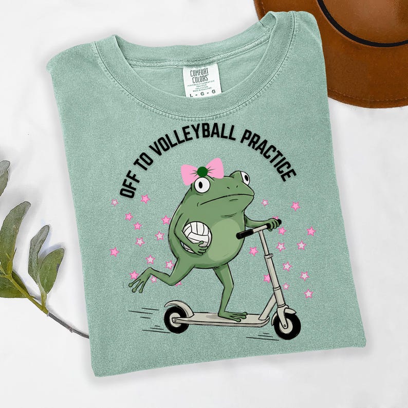 May include: A light green t-shirt with the text "OFF TO VOLLEYBALL PRACTICE". The shirt features a cartoon frog riding a scooter and holding a volleyball. The frog wears a pink bow and is surrounded by pink stars.