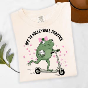 May include: Cream-colored t-shirt featuring a cartoon frog riding a scooter and holding a volleyball. The frog wears a pink bow and the text reads "OFF TO VOLLEYBALL PRACTICE" in a circular design with pink stars.