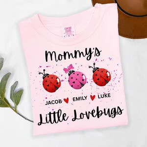 May include: Pink t-shirt with the words "Mommy's Little Lovebugs" and three ladybug graphics. The ladybugs are red and pink with the names Jacob, Emily, and Luke below them. The shirt has a Comfort Colors tag.