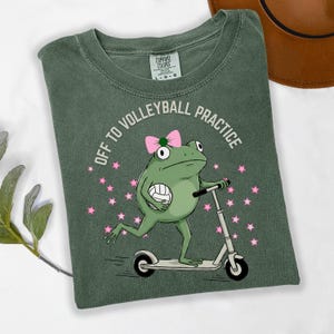 May include: Green t-shirt with a frog graphic riding a scooter and holding a volleyball. The text "OFF TO VOLLEYBALL PRACTICE" is printed above the frog. Pink stars are scattered around the frog.