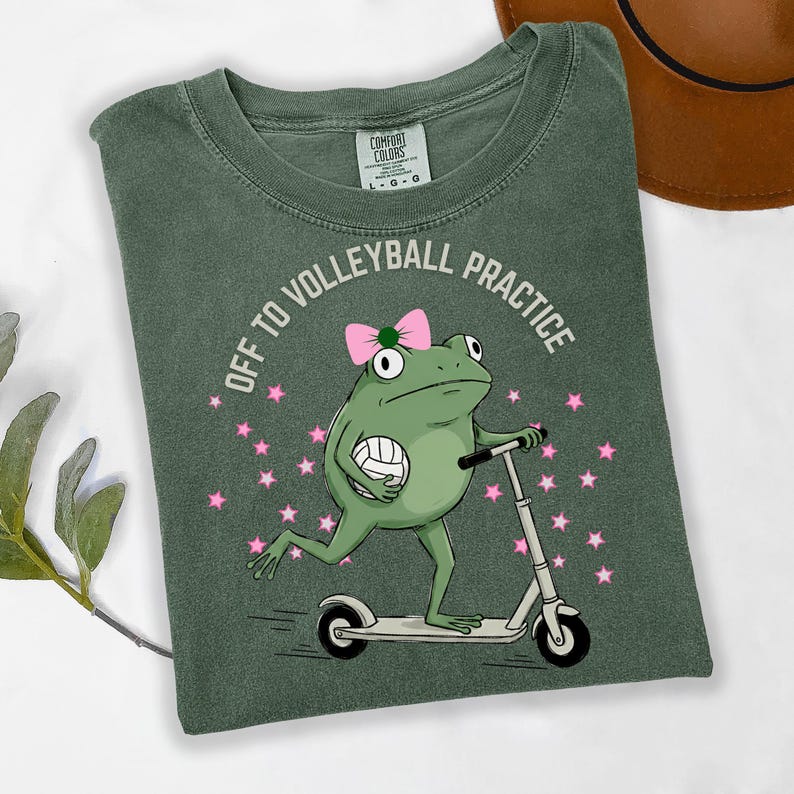 May include: Green t-shirt with a frog graphic riding a scooter and holding a volleyball. The text "OFF TO VOLLEYBALL PRACTICE" is printed above the frog. Pink stars are scattered around the frog.