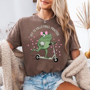 May include: Brown t-shirt with a graphic of a green frog riding a scooter and holding a volleyball. The text "OFF TO VOLLEYBALL PRACTICE" is printed around the frog. Pink stars surround the frog.