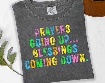 May include: A gray t-shirt with the text "PRAYERS GOING UP... BLESSINGS COMING DOWN" in colorful, distressed lettering. The shirt has a crew neck and a Comfort Colors tag. A brown hat is partially visible in the background.