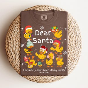 May include: Brown t-shirt with a festive design featuring yellow ducks wearing holiday accessories. The text "Dear Santa" is above the phrase "I definitely don't have all my ducks in a row!" A fun, seasonal graphic tee.