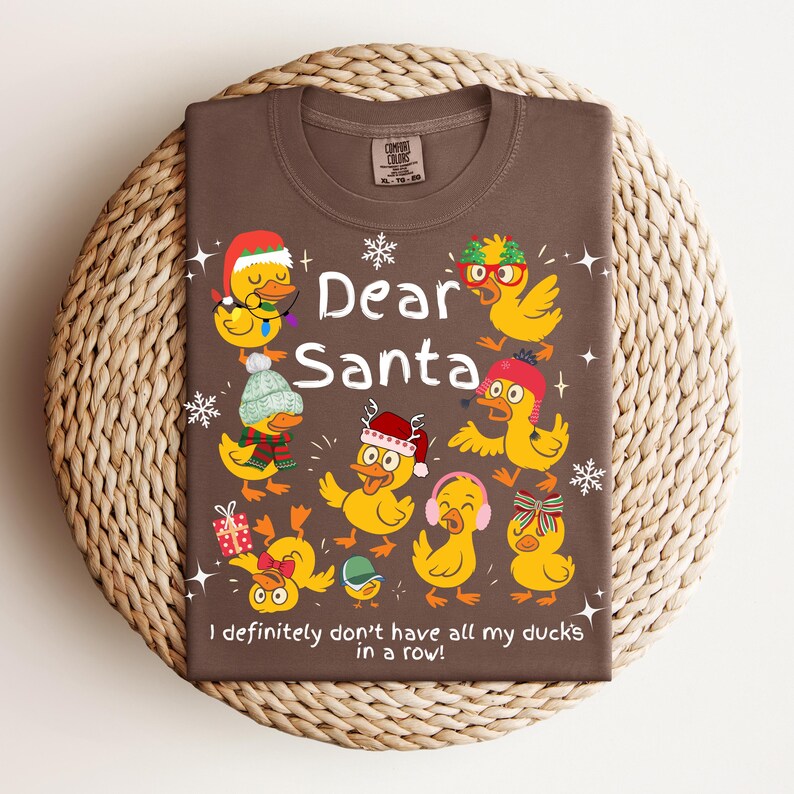 May include: Brown t-shirt with a festive design featuring yellow ducks wearing holiday accessories. The text "Dear Santa" is above the phrase "I definitely don't have all my ducks in a row!" A fun, seasonal graphic tee.