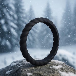 May include: A black, braided silicone ring rests on a snow-covered rock. The ring has a rope-like texture. The background shows a snowy forest with falling snowflakes, creating a winter scene.