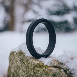 May include: A black silicone ring sits on a snow-covered rock. The ring is a solid, matte black with a smooth, rounded design. The background is a blurred winter scene with falling snow.