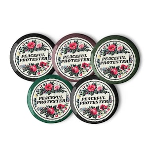 Op de afbeelding: Vijf ronde knopen met bloemmotieven en de tekst "PEACEFUL PROTESTER". De knopen hebben zwarte, bordeauxrode en groene randen. Het bloemmotief toont rode en roze rozen met groene bladeren.