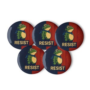 Puede incluir: Cinco botones redondos con una rana verde y amarilla con una bufanda azul. El fondo es rojo y azul, con la palabra "RESIST" en blanco.